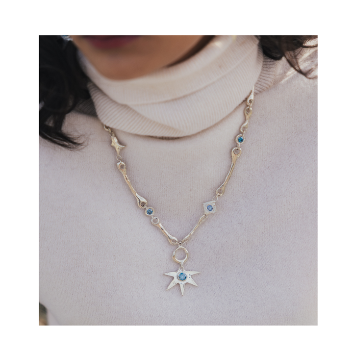 A person wearing a cream turtleneck sweater and the Madeleine Sculptural Jewellery Bone Necklace with Star Centre, featuring silver links, blue gemstones, and a large star-shaped pendant.