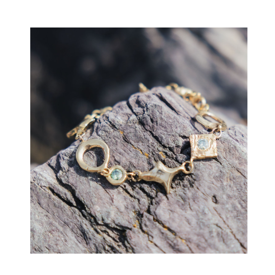 The Spark Bracelet by Madeleine Sculptural Jewellery, featuring various shaped charms and small blue gemstones, rests on a textured gray rock in natural sunlight.