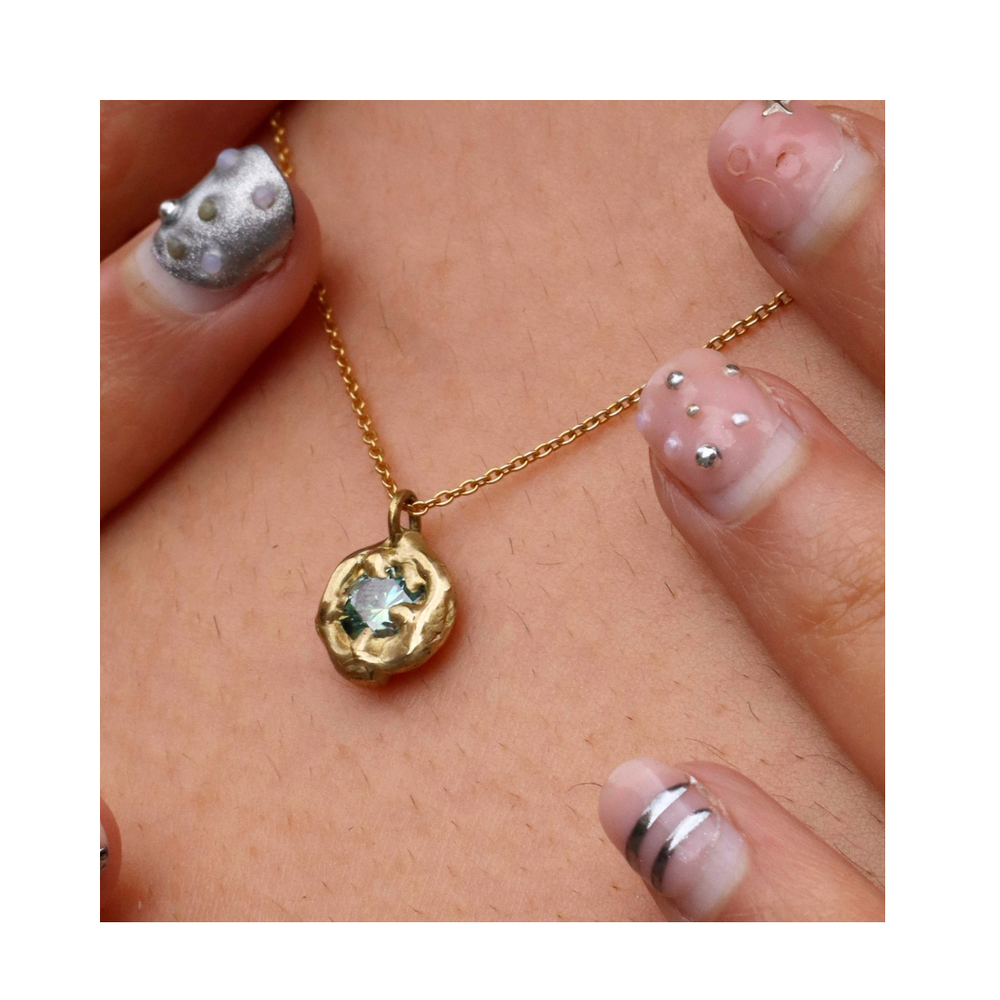 Close-up of hands with elegant, decorated nails holding the Molten Necklace with Moissanite by Madeleine Sculptural Jewellery—a hand-carved gold piece featuring a round, textured pendant and shimmering light blue moissanite.