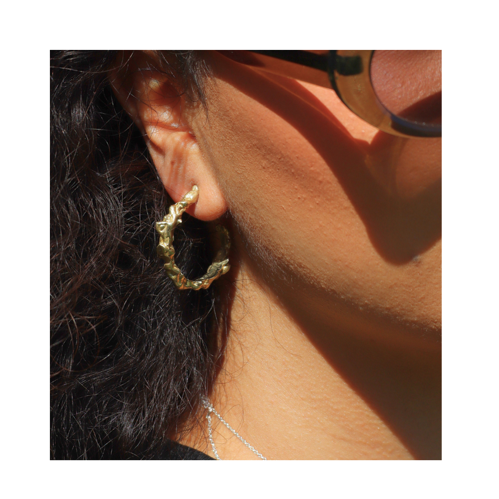Close-up of a person wearing Molten Hoops Large by Madeleine Sculptural Jewellery—textured gold earrings made from recycled brass. They have wavy dark hair, sunglasses, a delicate silver necklace, and warm lighting on their skin.