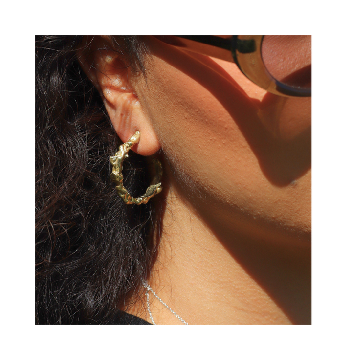 Close-up of a person wearing Molten Hoops Large by Madeleine Sculptural Jewellery—textured gold earrings made from recycled brass. They have wavy dark hair, sunglasses, a delicate silver necklace, and warm lighting on their skin.