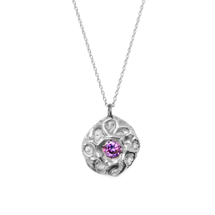 The Multi Stone Moissanite Silver Necklace by Madeleine Blaine is showcased on a white background, featuring a textured, round pendant adorned with a central purple gemstone and shimmering lab-grown moissanite stones, all suspended from a delicate silver chain.