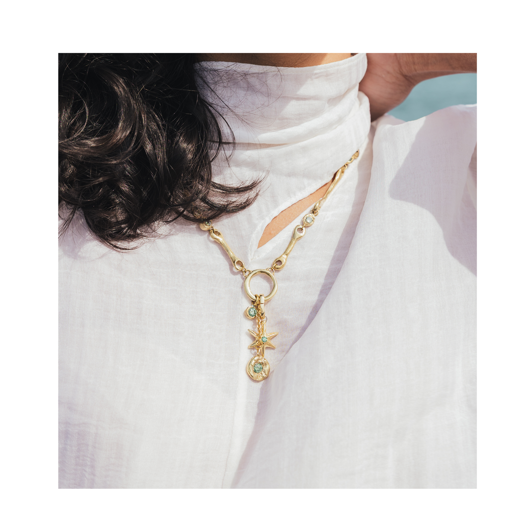 A person wearing a white high-neck blouse and the Lumen Necklace by Madeleine Sculptural Jewellery—featuring gold star-shaped pendants with a green gem—photographed from the shoulders up, with dark hair partially visible on one side.