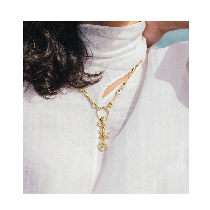 A person wearing a white high-neck top and the Madeleine Blaine Lumen Necklace—a gold handcrafted piece with a circular pendant featuring a star and a green gemstone—shown from the shoulders up.