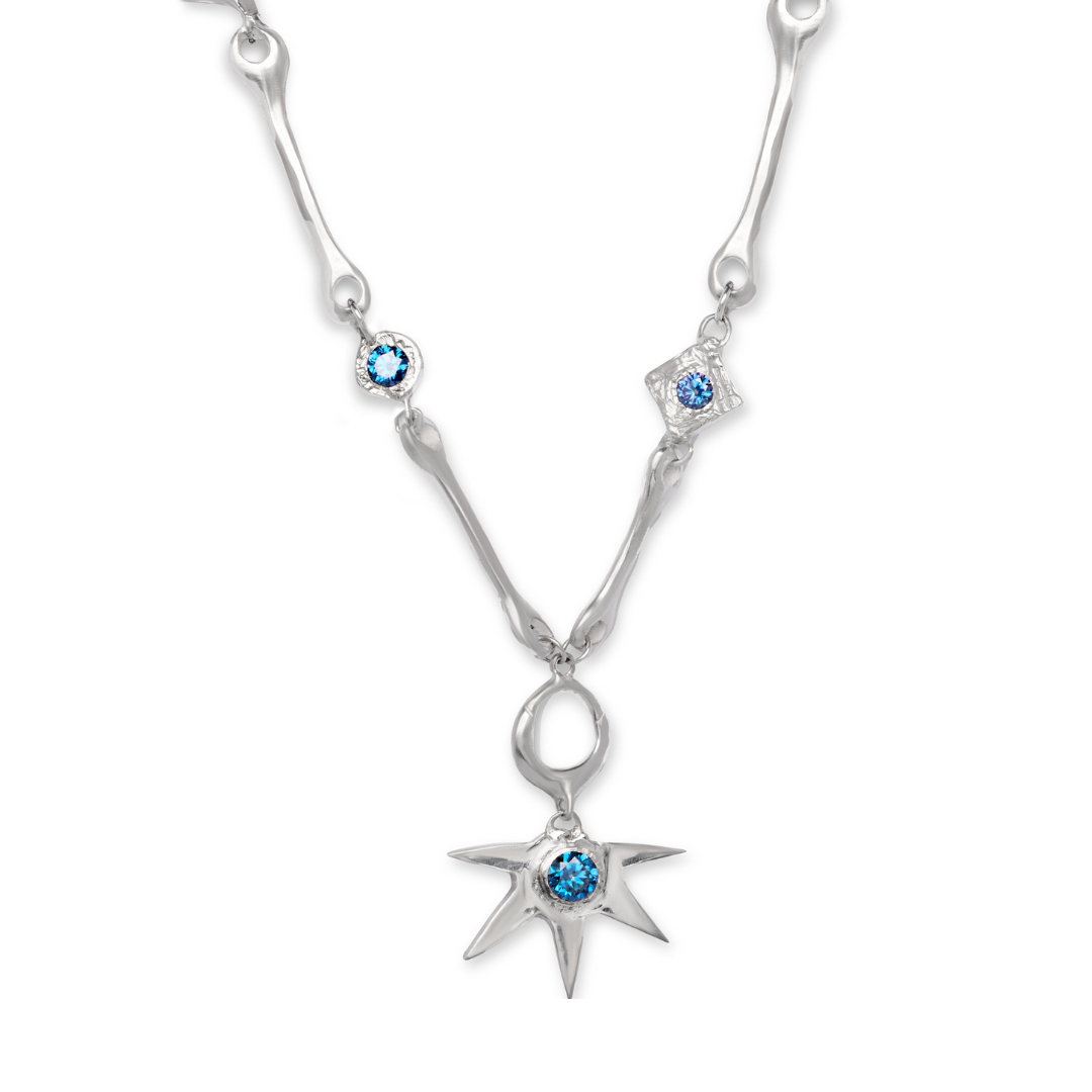 The Réalta Necklace with Moissanite by Madeleine Blaine features a silver chain adorned with blue gemstones, showcasing a star-shaped pendant and two smaller gemstone accents along the chain, set against a white background.