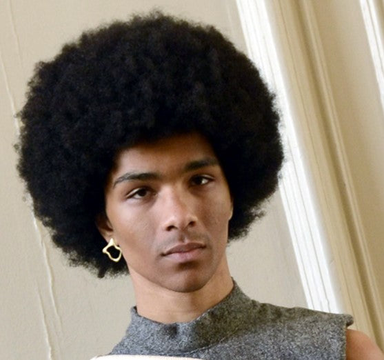A person with a large rounded afro wears a sleeveless, high-collared gray top and a gold Asymmetrical Molten Stud earring by Madeleine Sculptural Jewellery, standing indoors in front of a light-colored wall.