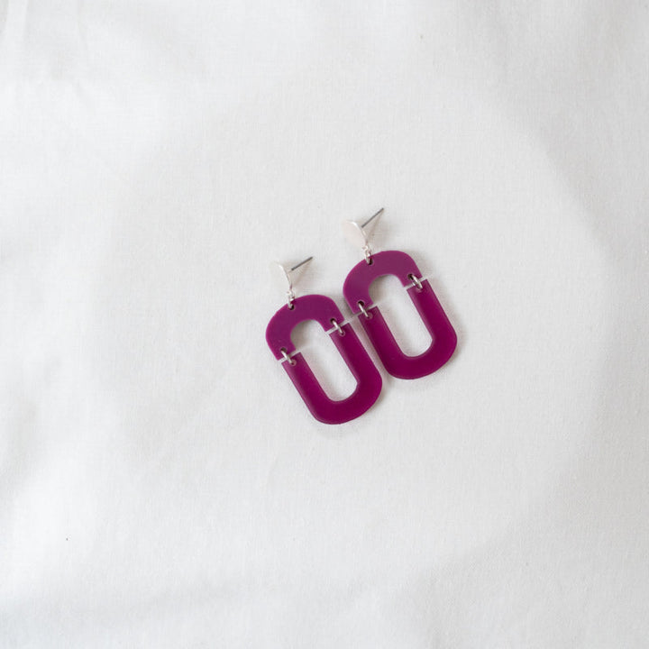 A pair of Magenta Oval Misty Acrylic Silver Earrings from Studio Nok Nok hang elegantly from silver-colored triangular studs on a white fabric background.