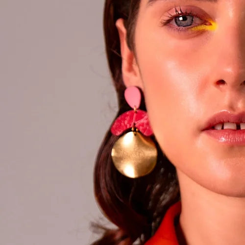Close-up of a woman with light skin wearing Shock of Grey's Galaxy Megadrop Earrings in Red and Pink, yellow eyeshadow on her inner eye corner, and an orange top. Only half of her face is shown.