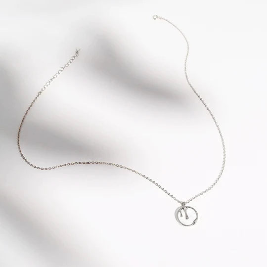 A delicate sterling silver necklace with a thin chain and a small round pendant featuring an abstract design rests on a white, textured surface. The minimalist style of the Melting Pendant Necklace Silver by Marie June Jewellery stands out against the soft, undulating background.