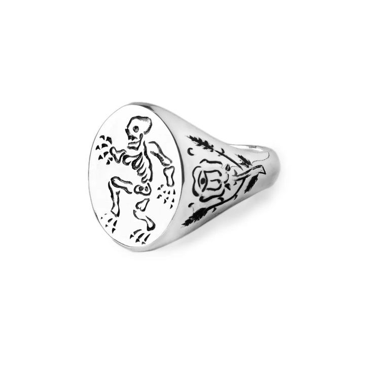 The Sterling Silver Memento Mori Signet Ring by Cosmic Boulevard features a skeleton and scattered dots on the face, plus a trident symbol and ornate detailing on the side.