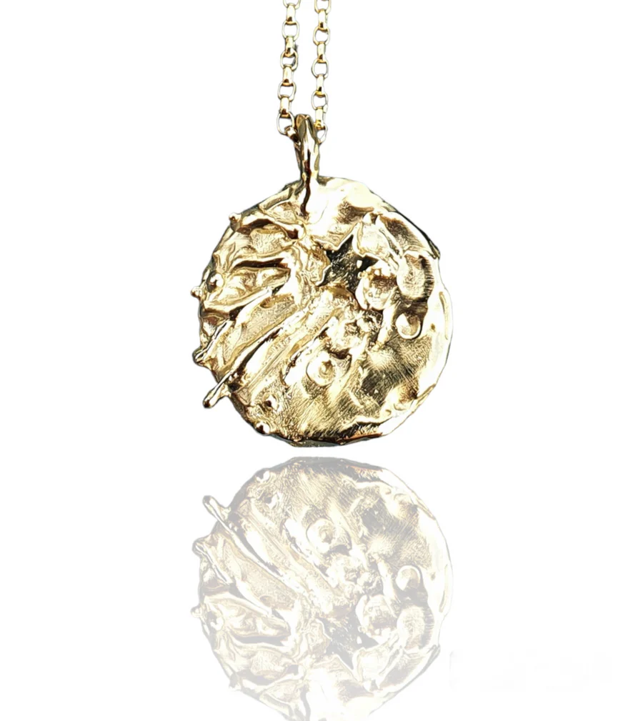 The Meteor at the Love Parade Gold Plated Necklace by Pale Raven features a round, textured coin pendant with a raised star on a delicate chain, beautifully reflected on a white surface.