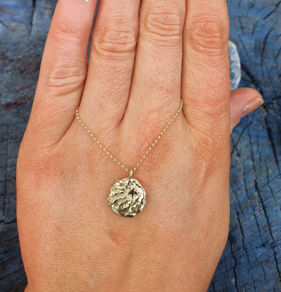 A hand with manicured nails displays the Pale Raven Meteor at the Love Parade Gold Plated Necklace, featuring a round, textured coin pendant, resting on the back of the hand against a dark blue wooden surface.