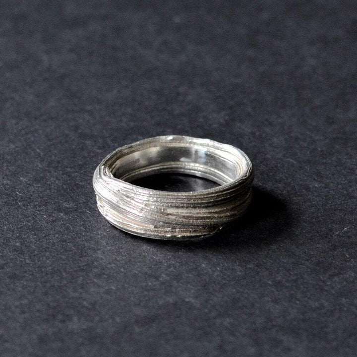 An intricate and rustic Mitsuro Silver Ring by Abbie Dixon lies on a dark, textured surface. The ring features multiple layers of thin, overlapping bands that create a unique, woven appearance, reminiscent of the Mitsuro Hikime method, adding depth and character to its design.