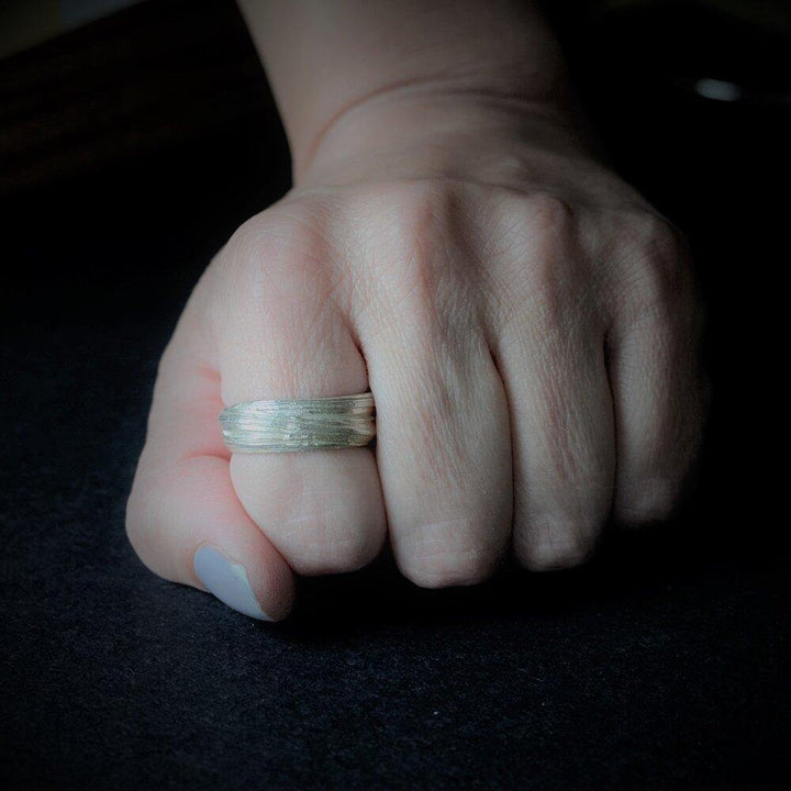 A close-up of a hand with a sterling silver ring on the middle finger. The hand is resting on a dark surface, and the fingers are slightly curled inward. The lighting focuses on the Mitsuro Silver Ring by Abbie Dixon, casting a shadow on the surface.