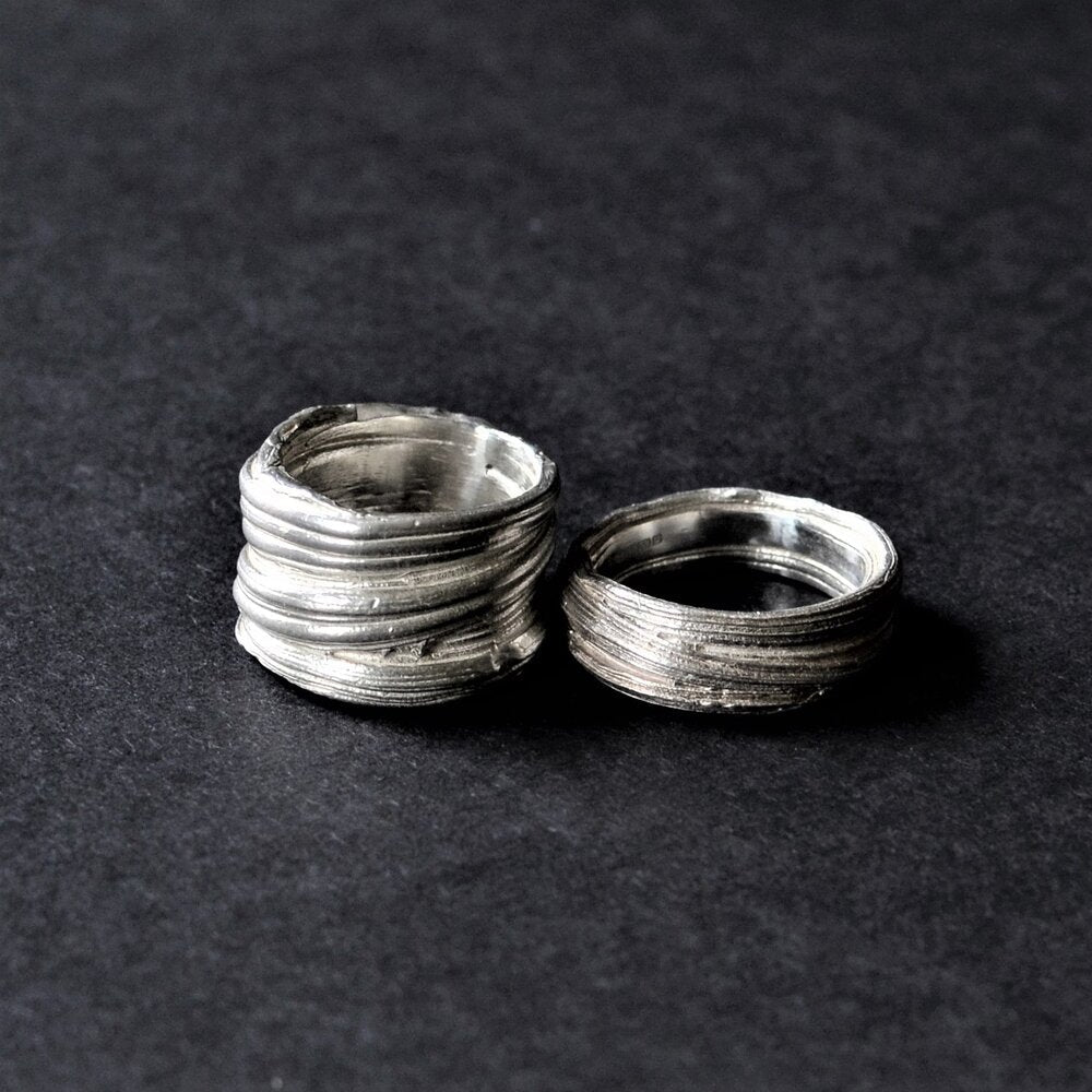 Two Mitsuro Silver Rings by Abbie Dixon with a rustic, textured finish are placed side by side on a dark, textured surface. One ring is slightly larger and has a more pronounced, layered design reminiscent of the lost wax method, while the other ring is smaller with a subtler texture.