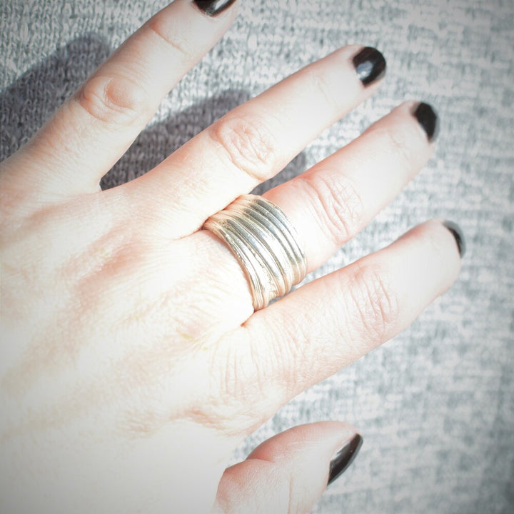 Close-up of a hand with black-painted nails, wearing multiple stacked metallic rings on the middle finger. Among them, an Abbie Dixon Mitsuro Silver Ring catches the eye. The background is a textured gray surface. The rings appear to be different shades of silver and gold, varying in thickness.