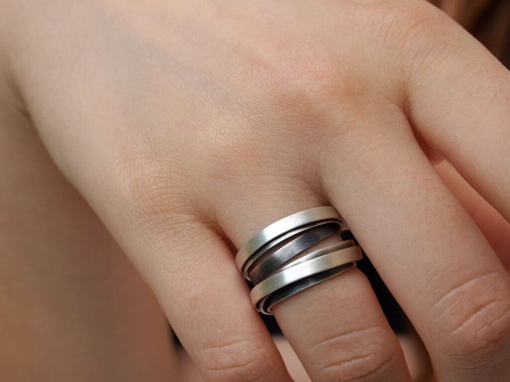 A close-up of a hand wearing three stacked Mo Silver and Oxidised Rings from MarCucurella on the middle finger. The rings, possibly crafted from handmade sterling silver, feature a simple, sleek design. The skin tone is light with a soft, smooth texture.