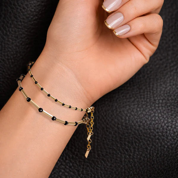 A manicured hand with gold-tipped French polish wears the Scribble & Stone Nightsky Linear Bracelet 3mm, featuring delicate gold and black beads, against a textured black background.