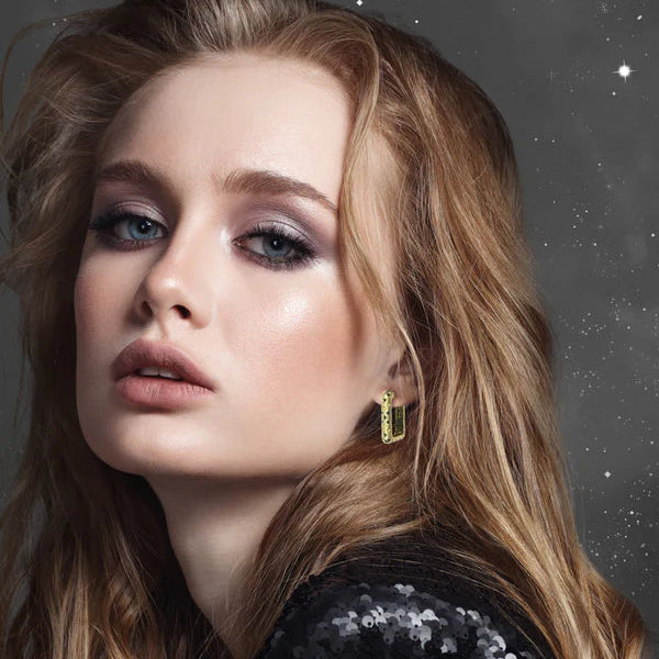 A woman with long, wavy blonde hair and blue eyes wears shimmery eye makeup, nude lipstick, a sparkling black top, and Scribble & Stone’s Starry Night Earrings, set against a starry night background.