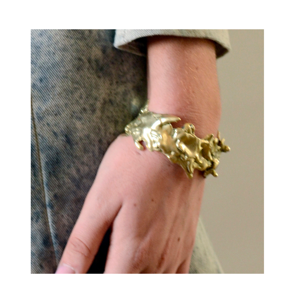 A hand rests casually by their side, adorned with the Molten Cuff by Madeleine Sculptural Jewellery—a striking, chunky gold bracelet that stands out against a grey textured outfit.