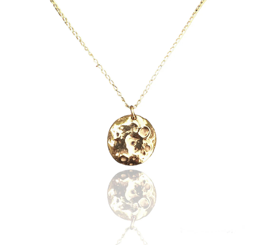The Pale Raven Molten Moon Gold Plated Necklace showcases a handcrafted round celestial pendant with a textured, moon-like surface, displayed on a white background with a subtle reflection underneath.