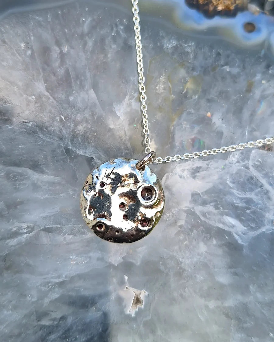 The Molten Moon Silver Necklace by Pale Raven features a handmade sterling silver pendant with crater-like dimples, suspended from a delicate chain and set against translucent, marbled stone.