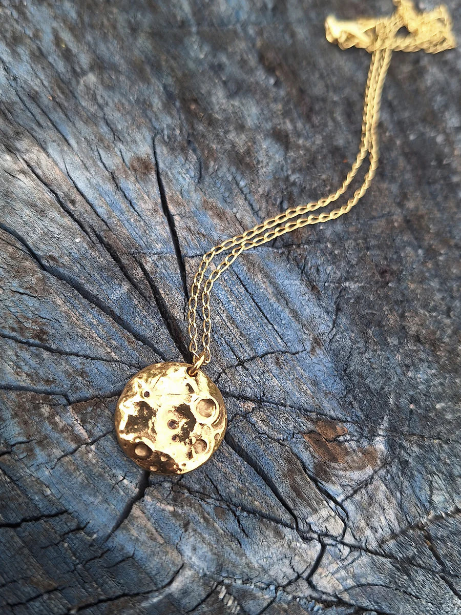 The Molten Moon Gold Plated Necklace by Pale Raven features a handcrafted, round celestial moon pendant and is displayed on weathered, cracked wood.