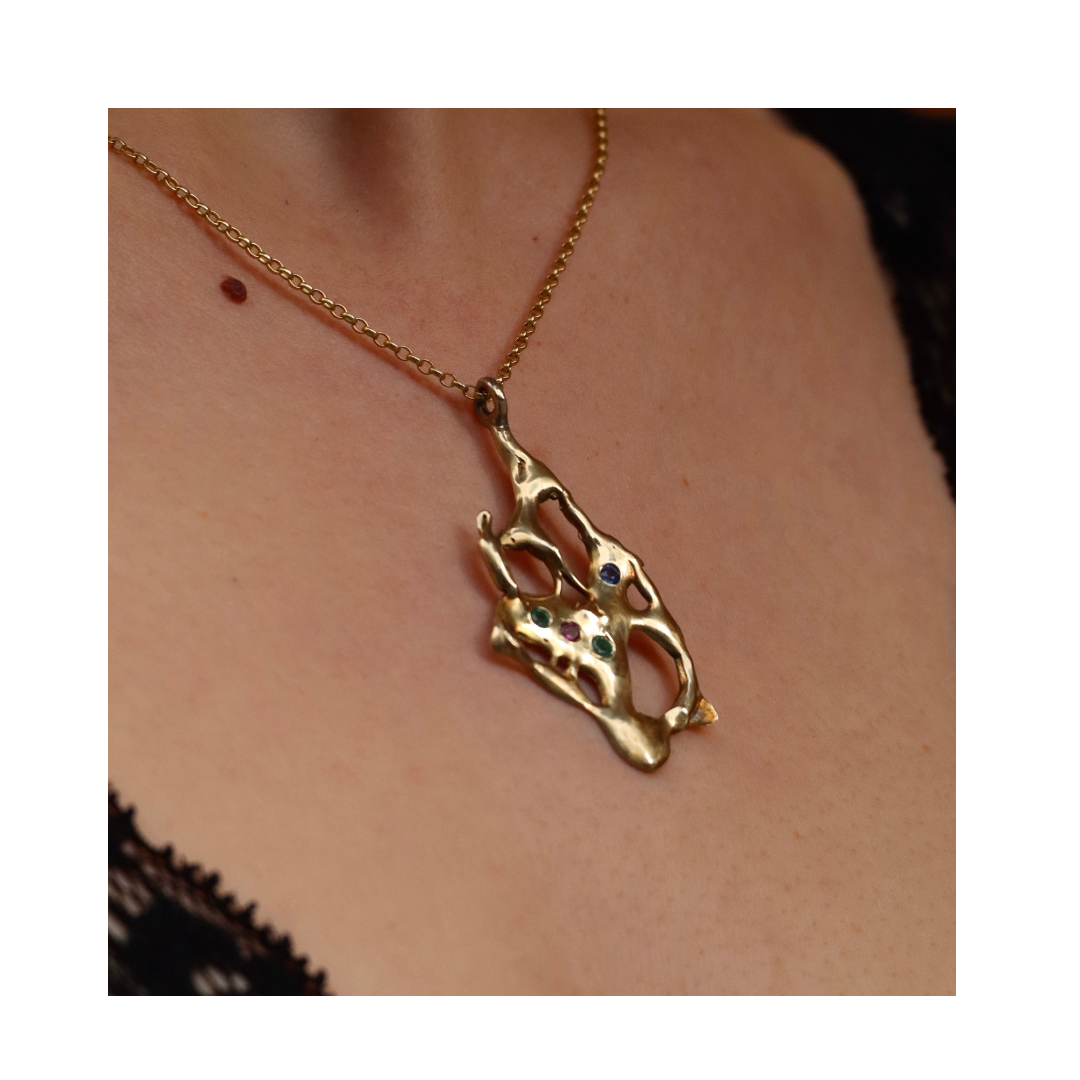 A person wearing the Madeleine Sculptural Jewellery Molten Pendant with Gemstones, crafted from recycled brass and accented with colorful stones, set against bare skin and partially visible black lace clothing.