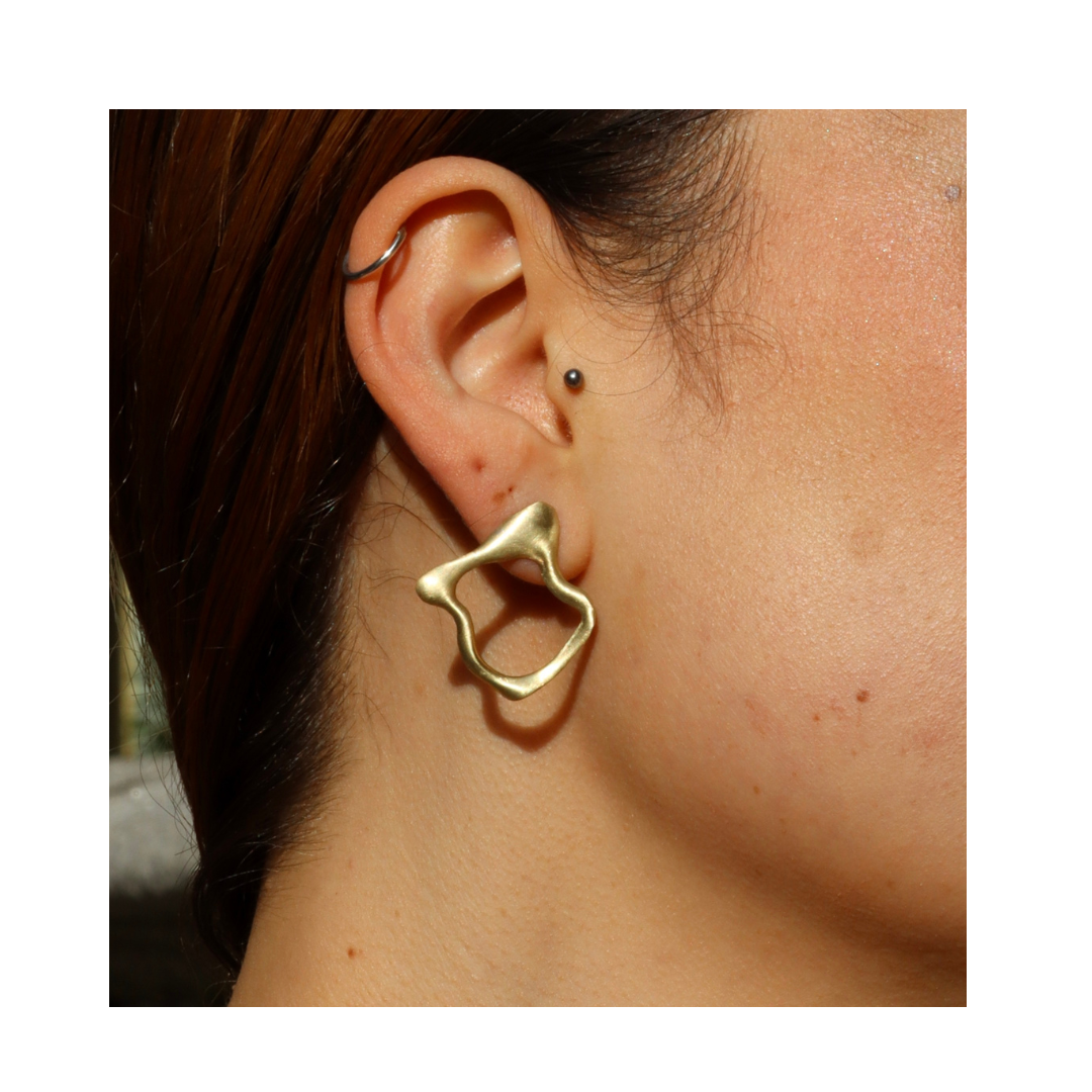 Close-up of a person’s ear wearing Madeleine Sculptural Jewellery’s Molten Studs—large, abstract-shaped gold earrings made from recycled brass—plus a small stud and thin hoop against a white background.