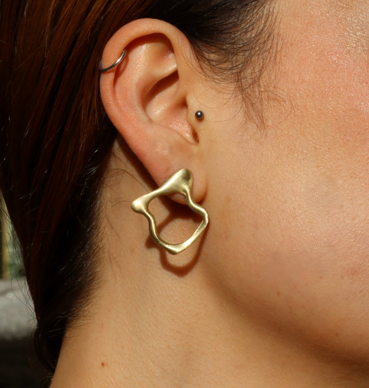 Close-up of a person's ear with short brown hair tucked behind it, showcasing Madeleine Sculptural Jewellery’s Asymmetrical Molten Studs—a large, irregularly shaped gold earring paired with a small hoop and tiny stud.