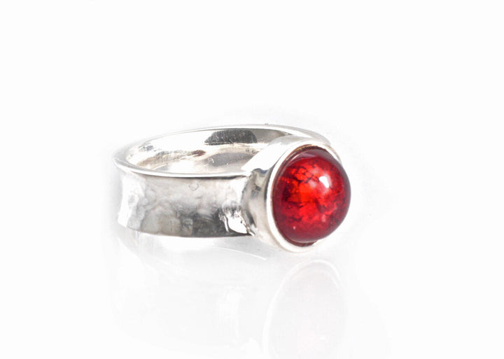 A sterling silver ring with a wide band, featuring a round, glossy red stone set in the center. The Murano Half Perle Silver Ring 10mm by Detlef Menard has a deep, vibrant hue and is smoothly polished. The band has a slightly hammered texture, giving it a unique and handcrafted appearance.