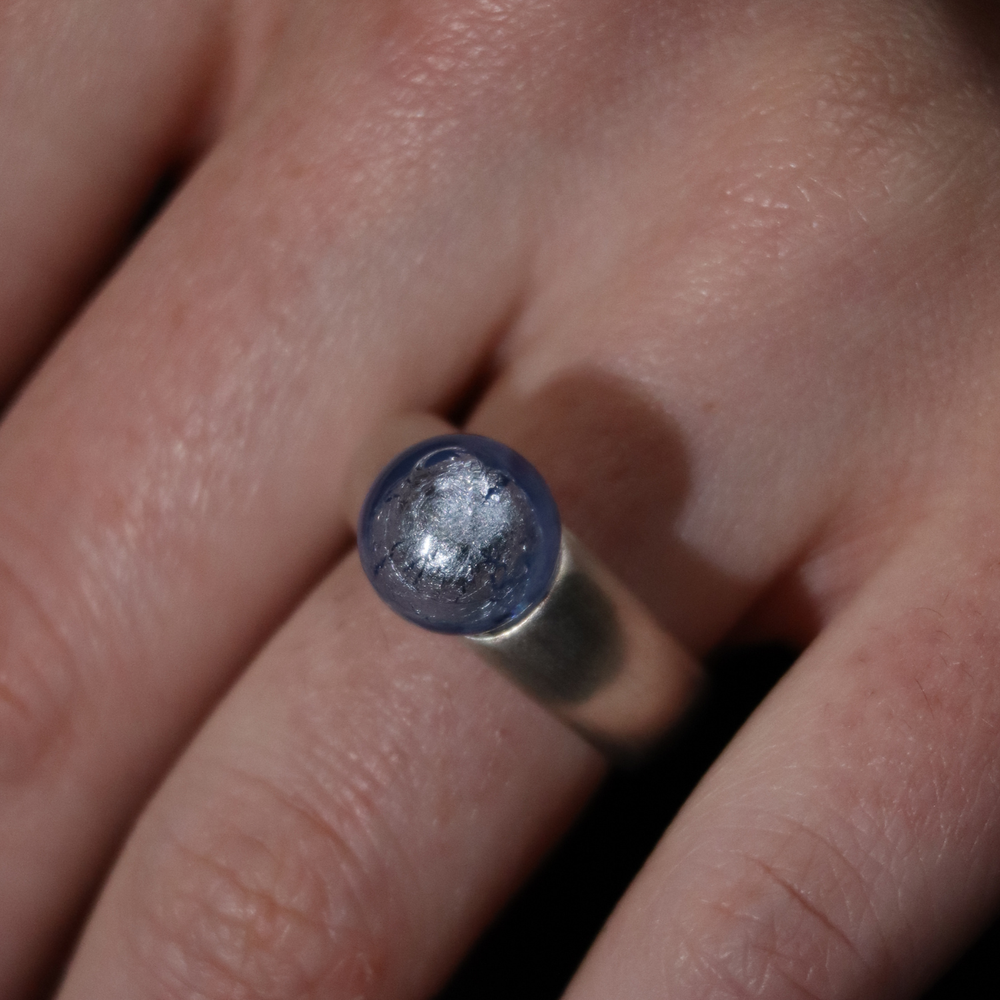 A close-up of a hand wearing the Detlef Menard Muranoperle Silver Ring 12mm, featuring a smooth, metallic finish and a round blue and silver marbled stone on top.
