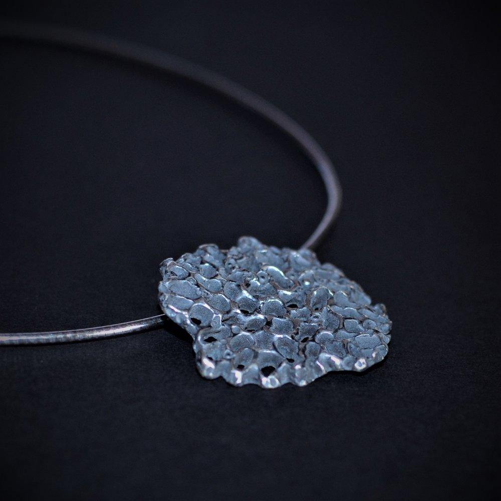 A close-up of a unique, irregularly shaped oxidised sterling silver pendant with a textured, pebble-like surface, hanging from a thin sterling silver omega chain. The dark background accentuates the intricate details of this piece of modern sculptural jewelry, the "Mythology Large Pendant Oxidised" by Abbie Dixon.