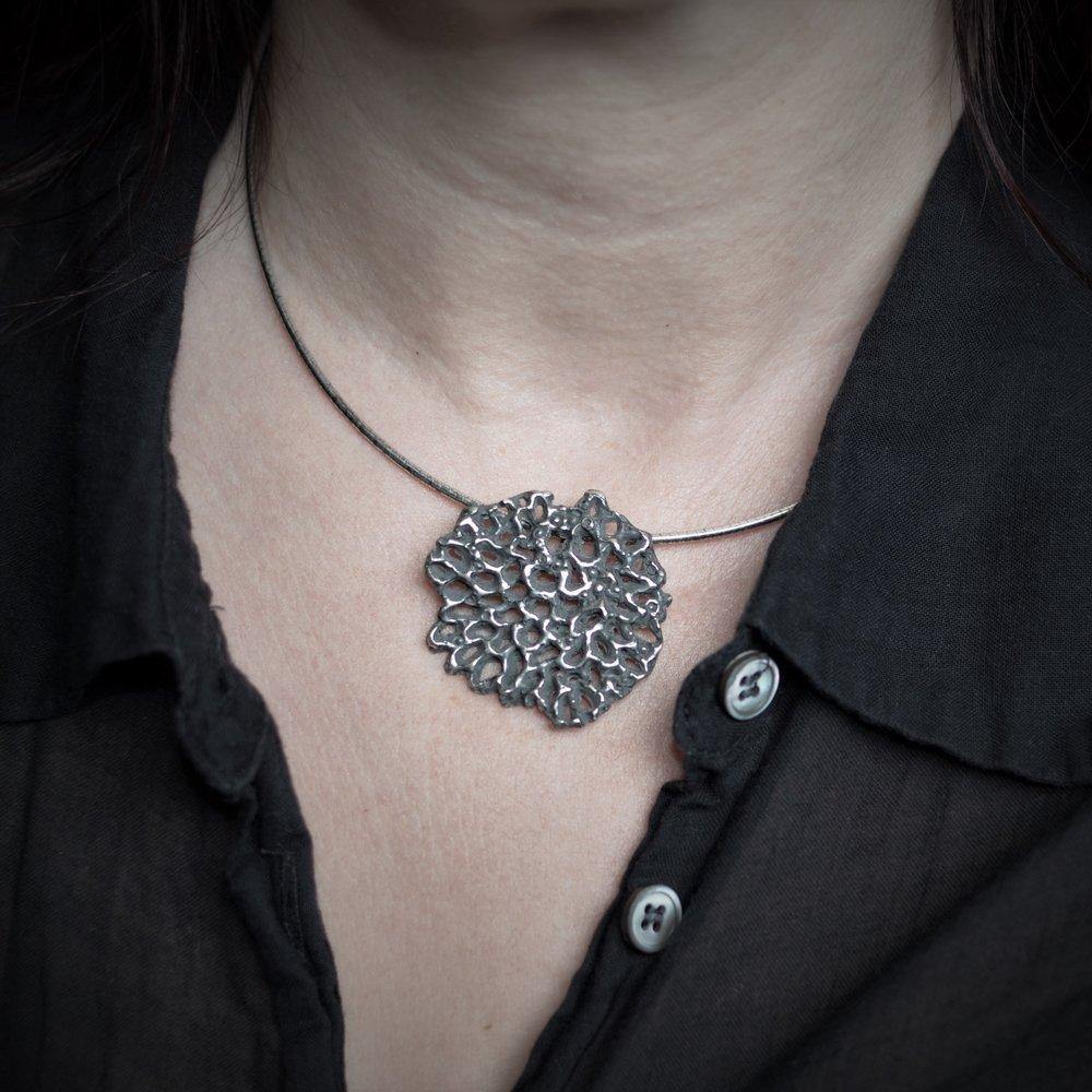 A person wearing a dark button-up shirt showcases an Abbie Dixon Mythology Large Pendant Oxidised with a textured, abstract flower design on a sterling silver omega chain. The image focuses on the neck and upper chest area, highlighting the intricate details of this modern sculptural jewelry piece.