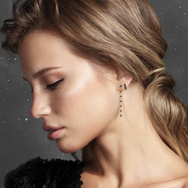 A woman with light brown hair styled back and natural makeup wears a black top and Scribble & Stone’s Nightsky Linear Earrings—14ct gold fill with small black spinel beads—against a gray, starry background.