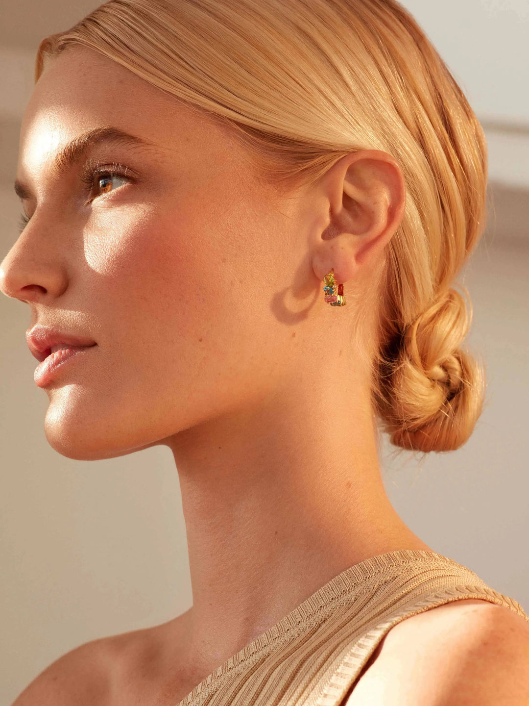 A woman with blonde hair in a low twisted bun wears a one-shoulder beige top and Shyla's Nila Huggies, looking to the left in warm natural light.