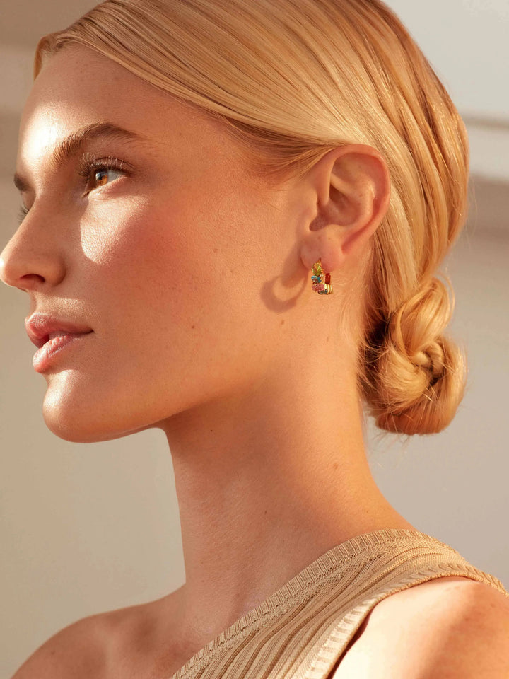 A woman with blonde hair in a low twisted bun wears a one-shoulder beige top and Shyla's Nila Huggies, looking to the left in warm natural light.