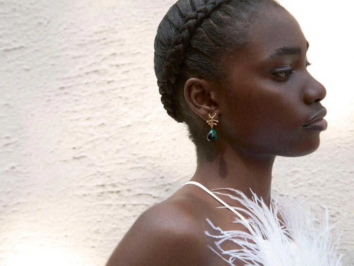 A person with dark skin and braided hair is seen in profile against a textured light background. They are wearing a white feathered outfit and a gold Shyla Nuria Earrings with a green gemstone. The lighting casts a soft glow, highlighting their serene expression.