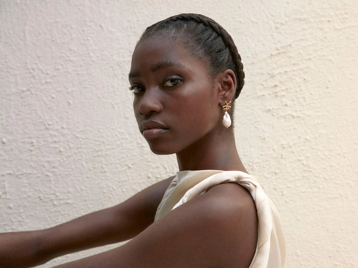 A person with dark skin wears a sleeveless, light-colored top and a pair of Shyla Nuria Earrings with a Baroque Pearl. They have their hair styled in braids and stand against a textured white wall, looking over their shoulder at the camera.