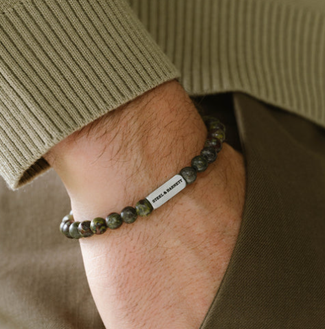 A close-up of a wrist wearing the Steel and Barnett Natural Ned 6mm Round Gemstone Bracelet Dragon Blood, featuring earthy tones and a metallic tag engraved with “STYLE & DIRECTION,” contrasting beautifully with a greenish-beige ribbed sweater.