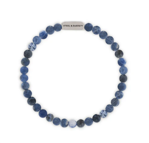 The Natural Ned 6mm Round Gemstone Bracelet by Steel and Barnett features matt sodalite beads in varied blue tones and a silver tag, offering an earthy and classic accessory style.