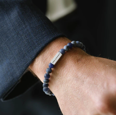 Close-up of a wrist wearing the Steel and Barnett Natural Ned 6mm Round Gemstone Bracelet in Matt Sodalite, featuring a small rectangular metal tag. This bracelet adds subtle elegance to earthy accessories, paired with a dark suit jacket.