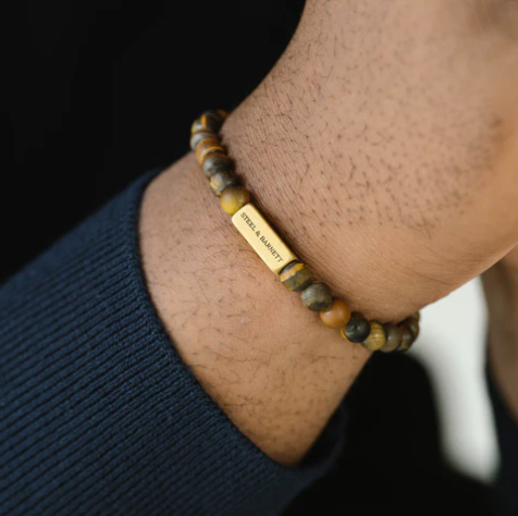 A close-up of a person's wrist wearing the Natural Ned 6mm Round Gemstone Bracelet in Matt Tiger Eye by Steel and Barnett, featuring a gold rectangular plate engraved with the brand name. The person is dressed in a navy blue long-sleeve shirt.