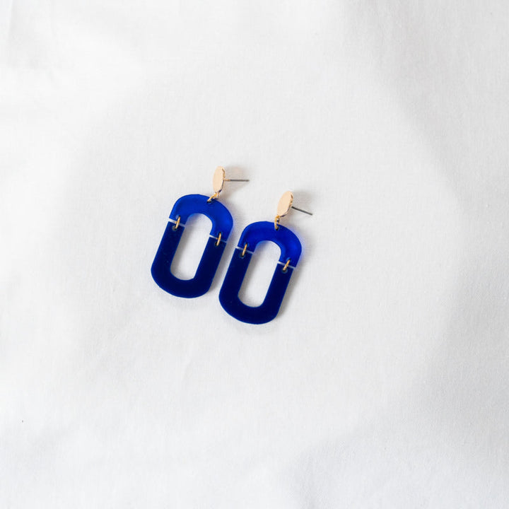 A pair of Navy & Cobalt Blue Acrylic Fire Earrings from Studio Nok Nok with matte blue hoops and small gold-plated brass studs lying on a white fabric background. The handmade earrings have a minimalist design with a modern aesthetic.