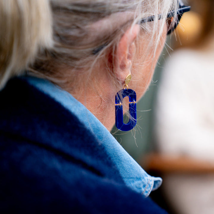 Close-up of a person with blonde hair tied back, wearing large, rectangular handmade earrings made from recycled acrylic and dark-rimmed glasses. The individual is dressed in a blue denim shirt and a dark jacket, with the focus primarily on their ear and the Navy & Cobalt Blue Acrylic Fire Earrings by Studio Nok Nok.