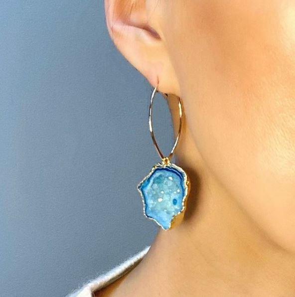 Close-up of a person wearing Aria V Blue Nebula Earrings featuring a vibrant blue geode agate slice pendant with specks of white. The earring has a golden trim and is handmade in Ireland, shown against a blurred gray background. Only the lower half of the individual's face and ear are visible.