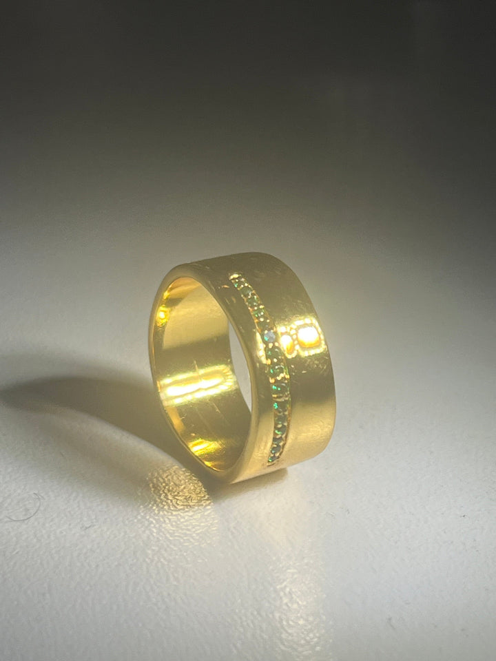 A gold chunky band ring with a sleek, wide profile sits on a smooth surface. The Shyla Nobu Ring features a horizontal line of small, green gemstones embedded along the center, adding a touch of elegance and sparkle to its design. The lighting casts a soft shadow, highlighting the ring's shine.