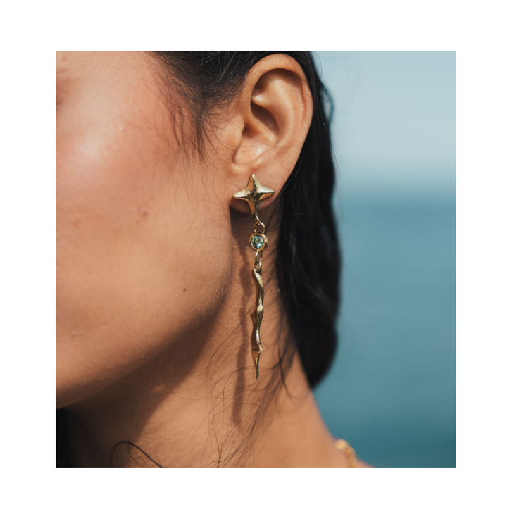 A close-up shot highlights a woman’s ear adorned with the Madeleine Blaine Nova Earrings Small with Moissanite: elegant, long, twisted gold earrings featuring a sparkling moissanite gemstone. Her dark hair is swept behind her ear, set against a softly blurred blue background reminiscent of the sea or sky.