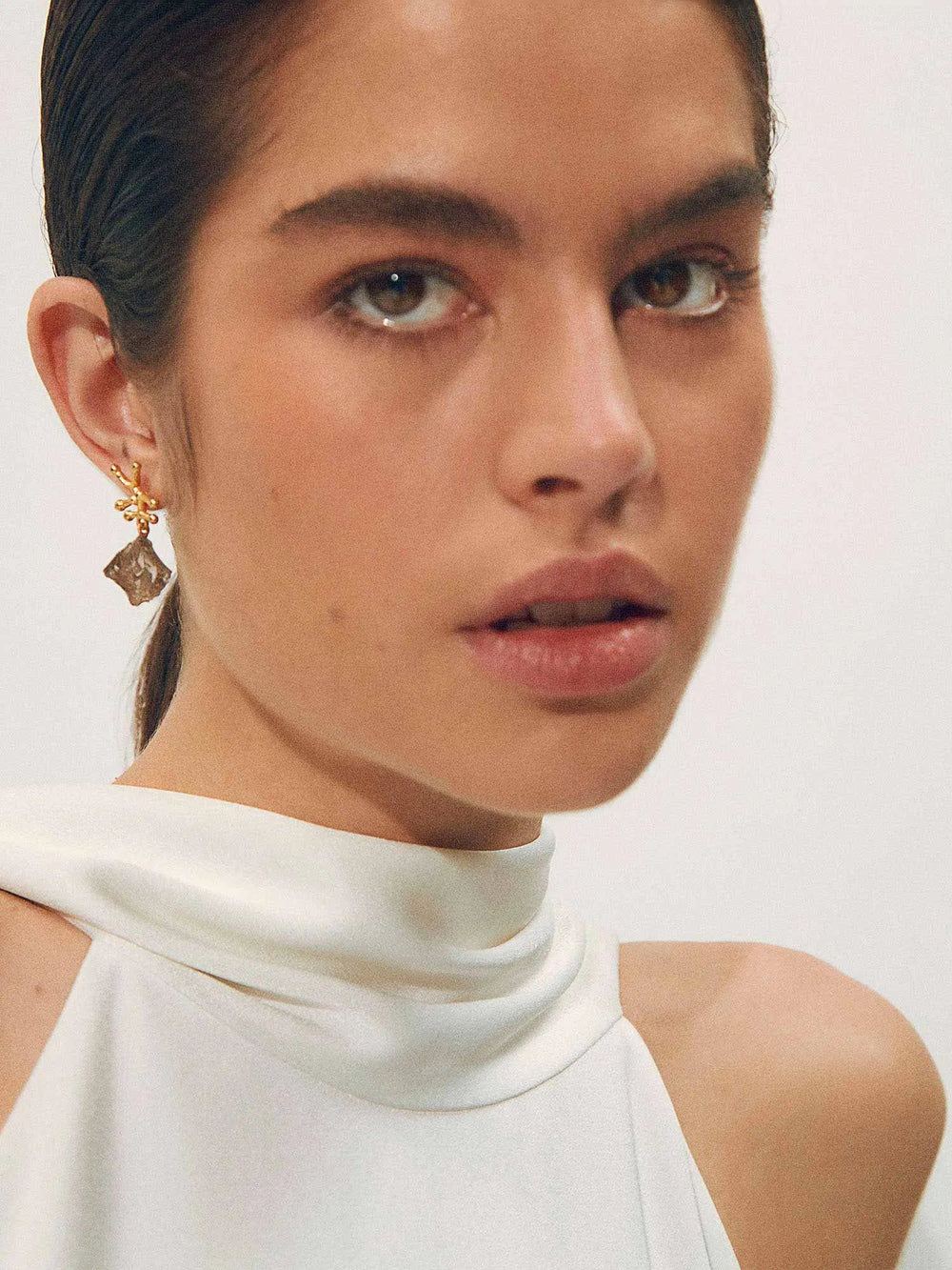 A woman with fair skin and striking eyes is shown from the shoulders up. She has dark, slicked-back hair and is wearing a sleeveless, high-neck white top. A gold Shyla Nuria Earring with a dangling Raw Smoky Crystal adorns her ear. The background is neutral, keeping focus on her face.