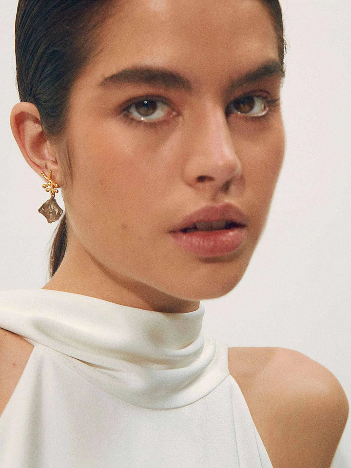 A woman with fair skin and striking eyes is shown from the shoulders up. She has dark, slicked-back hair and is wearing a sleeveless, high-neck white top. A gold Shyla Nuria Earring with a dangling Raw Smoky Crystal adorns her ear. The background is neutral, keeping focus on her face.